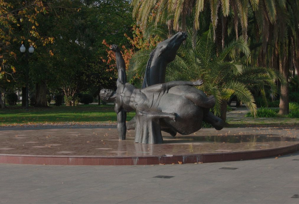 Large dark bronze sculpture of a man falling backwards beside a horse, on a round paved platform in a park with palm trees.