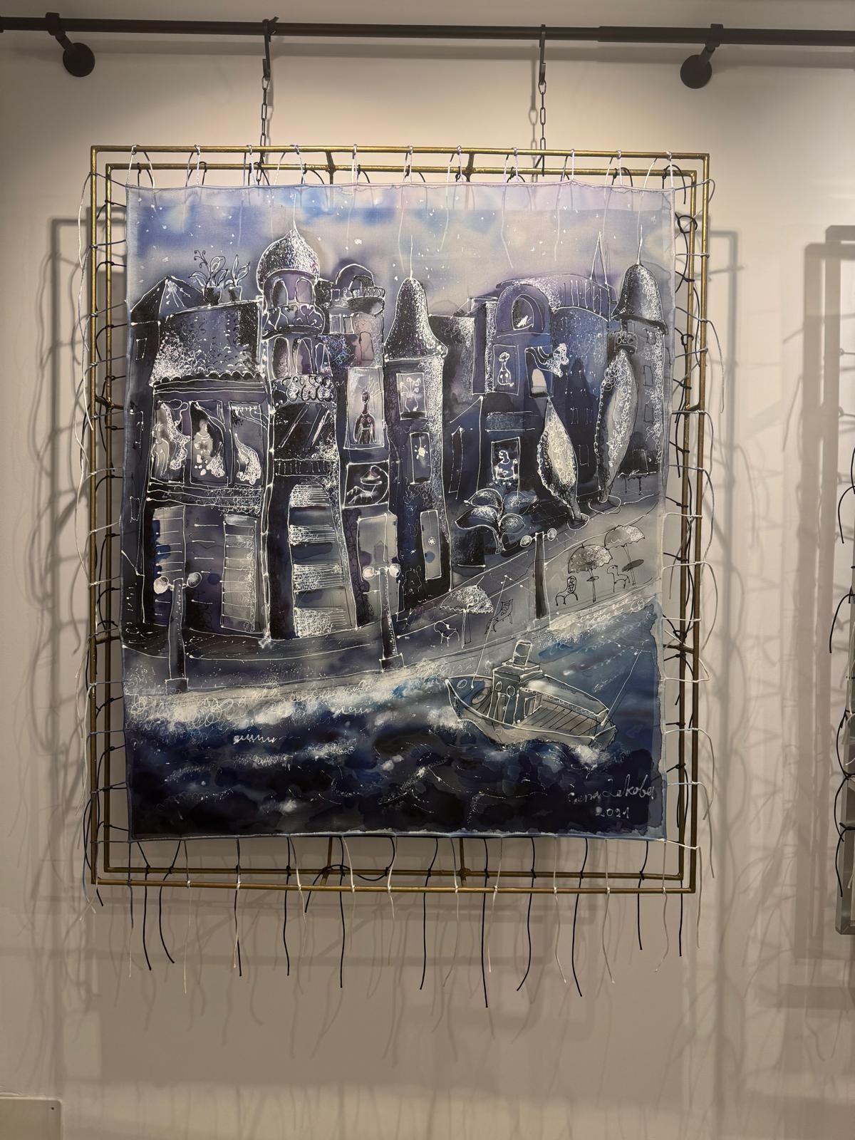 Silk painting in deep blues and white, stretched with cords inside a brass frame: a night embankment with tall domed and turreted houses, lit windows, street lamps and café umbrellas, and a small boat on dark waves in the foreground.